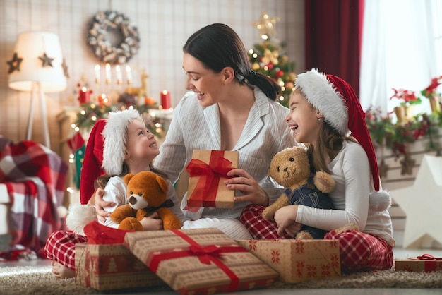 ¡Feliz Navidad y Felices Fiestas! Mamá alegre y sus lindas hijas intercambian regalos. Padres y niños pequeños divirtiéndose cerca del árbol en el interior. Familia cariñosa con regalos en la habitación.