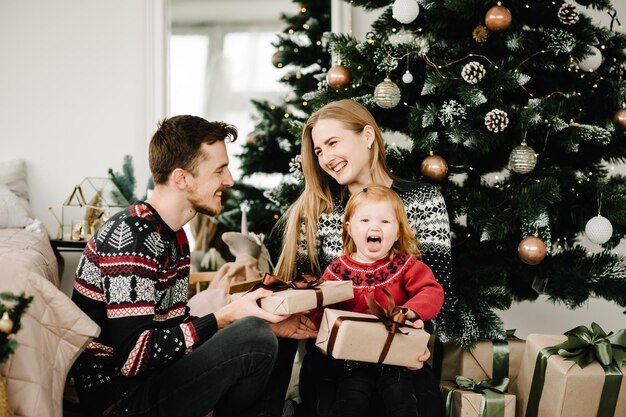 Feliz Navidad y Felices Fiestas Mamá papá le da un regalo a su hija cerca del árbol de Navidad Madre alegre padre abrazando a una linda niña juntos en casa Feliz Año Nuevo 2021