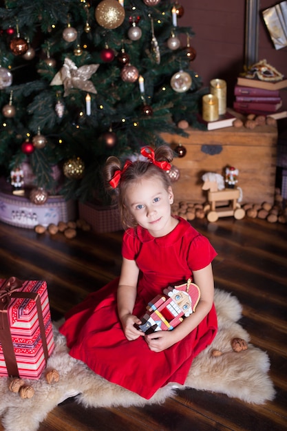 . Feliz Navidad, felices fiestas. Niña en un vestido rojo sostiene un cascanueces de madera vintage juguete cerca de un clásico árbol de Navidad en casa. Bailarina con el Cascanueces en la víspera de la superficie.