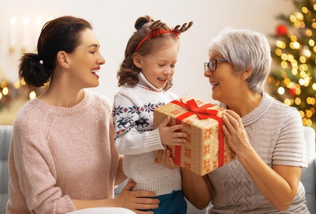 ¡Feliz Navidad y Felices Fiestas! Niño alegre que presenta regalos a mamá y abuela. Los padres y el niño se divierten cerca del árbol en el interior. Familia cariñosa con regalos en la habitación.