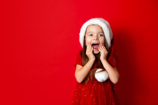 Feliz Navidad y felices fiestas Retrato de niña emocional en una gorra de Santa sobre un fondo rojo.