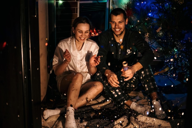 Feliz Navidad y Feliz Año Nuevo Una pareja joven y enamorada celebra la Navidad o la víspera de Año Nuevo en casa Una pareja romántica en pijama divirtiéndose con confeti en casa