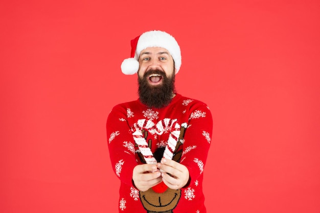 Feliz Navidad. feliz hombre barbudo con sombrero de santa claus. hipster brutal en suéter de Navidad de punto de invierno. chico maduro con palo de caramelo de vacaciones sobre fondo rojo. feliz Año Nuevo.