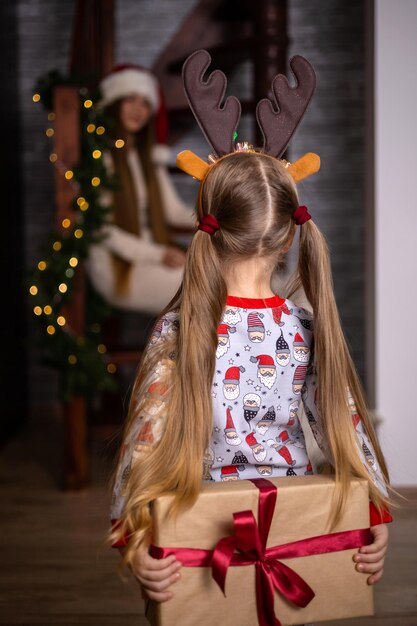 Feliz Navidad. Una hija de cabello largo le da a su madre un regalo escondido a sus espaldas en una caja con una cinta roja. Acogedor ambiente festivo de la casa. Foto de alta calidad