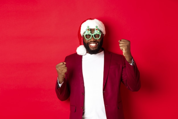 Feliz Navidad. Hombre negro alegre con gafas de fiesta divertidas y gorro de Papá Noel, sonriendo alegre, celebrando las vacaciones de invierno, de pie sobre fondo rojo.