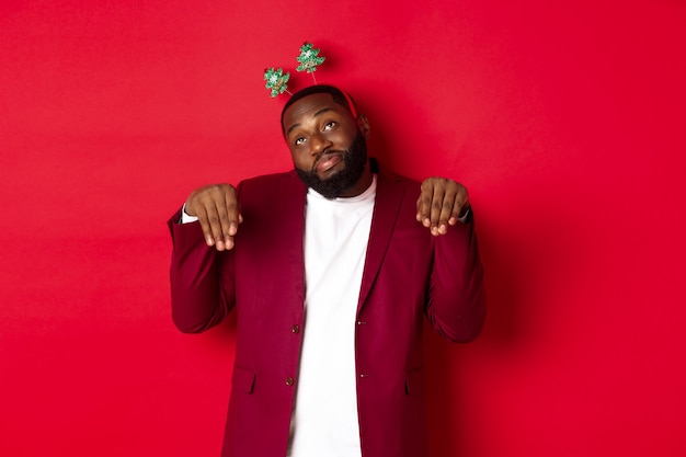 Feliz Navidad. Hombre negro tonto y divertido en diadema de fiesta, imitando conejito o lindo cachorro, de pie sobre fondo rojo.