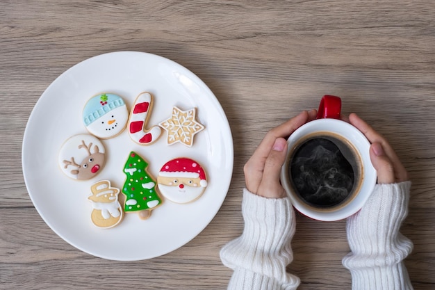 Feliz Navidad con mano de mujer sosteniendo la taza de café y galletas caseras en la mesa. Víspera de Navidad, fiesta, vacaciones y feliz año nuevo concepto