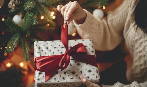 Feliz Navidad Manos en suéter abriendo regalo de navidad con lazo rojo en el árbol de navidad con luz