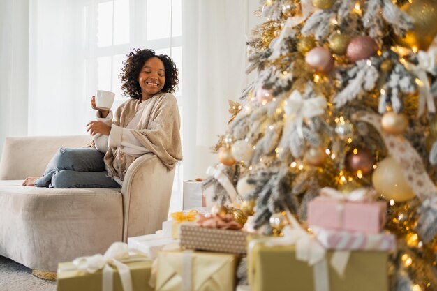 Feliz Navidad mujer afroamericana con una taza de bebida caliente café té sentado cerca del árbol de Navidad