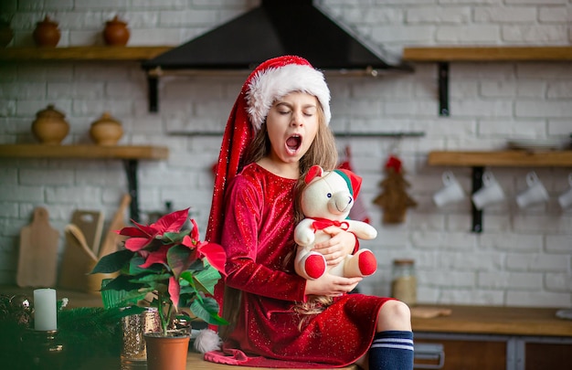 Feliz Navidad Niña linda con gorro de Papá Noel está sosteniendo un oso de peluche sentado en la cocina