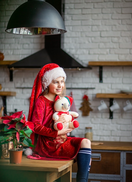 Feliz Navidad. Niña linda con sombrero de Santa está sosteniendo un oso de peluche sentado en la cocina, esperando las vacaciones.