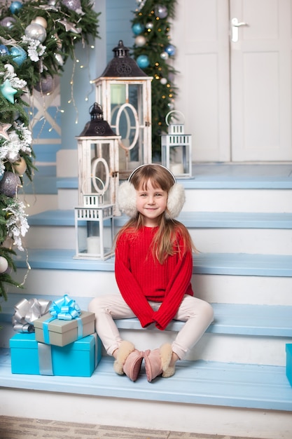 ¡Feliz Navidad! niña en suéter con regalo se sienta en el porche de la casa. El niño se sienta en la terraza decorada con adornos navideños y lámparas. niño abre regalo de navidad