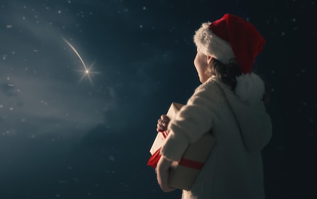 ¡Feliz Navidad! Niño lindo con regalo de Navidad. Niño feliz está pidiendo deseos mirando la estrella fugaz. Retrato de niña con regalo sobre fondo de cielo oscuro.