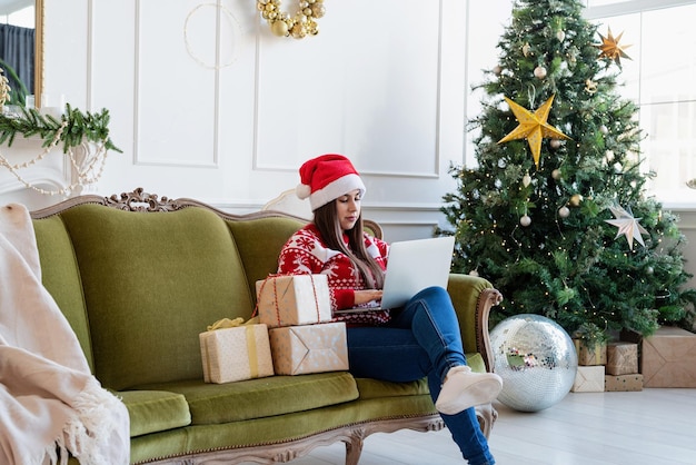 Feliz navidad y próspero año nuevo. Joven morena con gorro de Papá Noel sentado en el sofá verde trabajando en un portátil en una sala de estar decorada