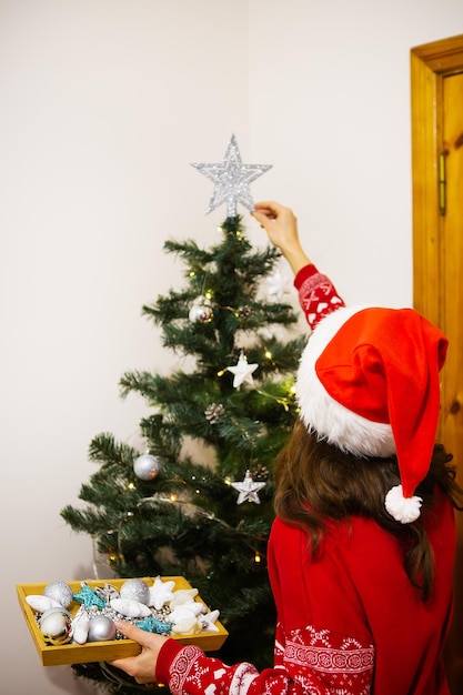 Feliz Navidad y Próspero Año Nuevo Las manos femeninas decoran el árbol de Navidad con bolas y juguetes Una mujer con un sombrero de Año Nuevo todo en rojo