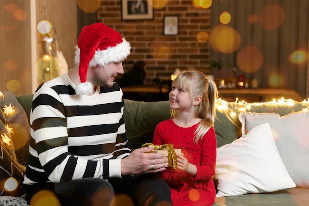 Feliz navidad y próspero año nuevo. Regalo sorpresa. Una pequeña hija sonriente feliz abre un regalo de Navidad con asombro. El padre le da a su hija un regalo en el interior de una casa decorada de forma festiva