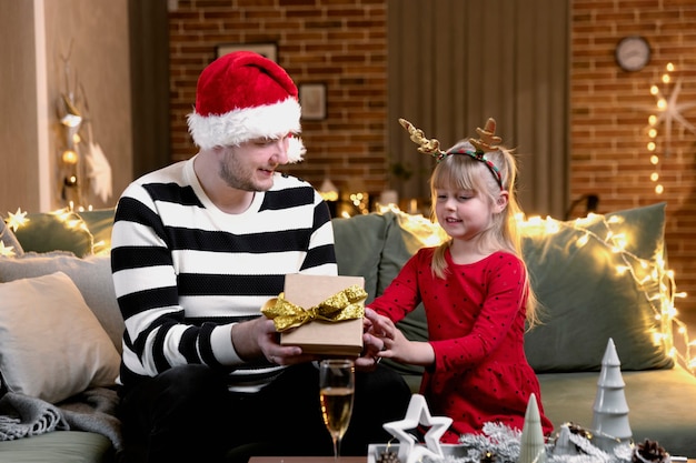 Feliz navidad y próspero año nuevo. Sorpresa de regalo de Navidad. Una pequeña hija sonriente feliz abre un regalo. Padre sosteniendo y dando giftbox en el interior de la casa con adornos navideños