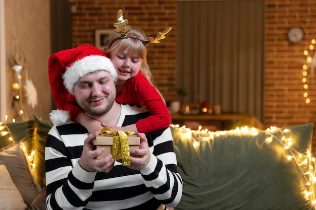 Feliz navidad y próspero año nuevo. Sorpresa de regalo de Navidad. Una pequeña niña sonriente hija abrazando al padre sosteniendo un regalo en el interior en casa decorada para las vacaciones. Hombre guapo tiene caja de regalo