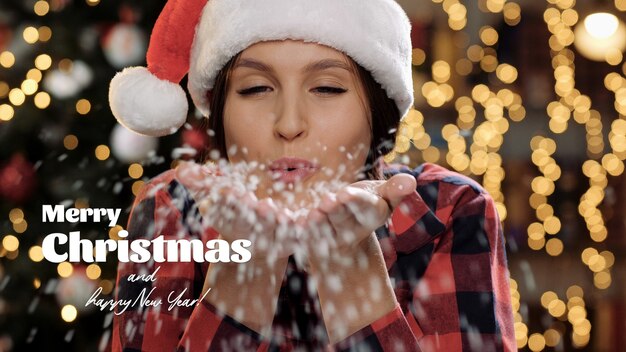 Foto feliz navidad y próspero año nuevo texto. hermosa mujer sonríe y sopla nieve blanca de sus palmas directamente a la cámara. de cerca