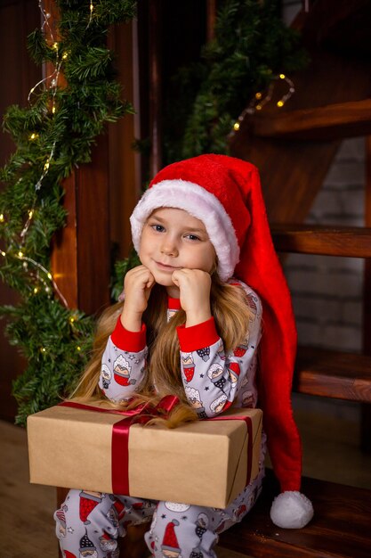 Feliz Navidad. Retrato de una linda chica con gorro de Papá Noel con una caja de regalo en las escaleras decoradas para Navidad. Foto de alta calidad