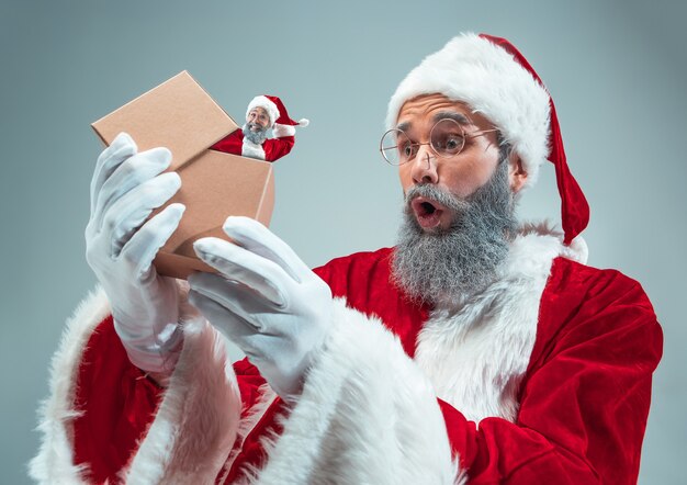 Feliz Navidad Santa Claus sobre fondo gris de estudio. Modelo masculino caucásico en traje tradicional de fiesta. Concepto de vacaciones, año nuevo, humor de invierno, regalos. Desempacando regalos, se preguntó.