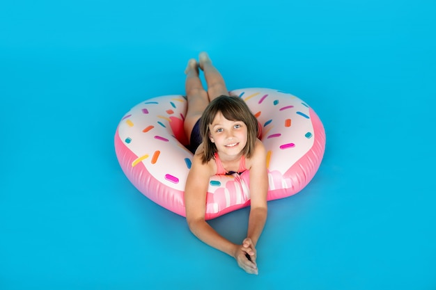 Feliz niña de 10 años con sombrero de paja en traje de baño con anillo de natación donut en una pared azul de color