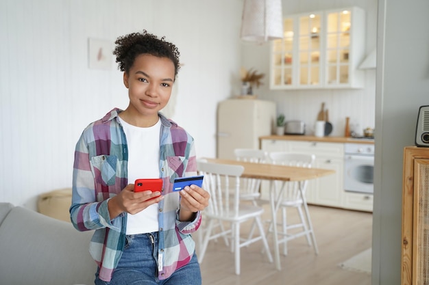 Feliz niña afroamericana está comprando en línea con teléfono móvil y tarjeta de crédito desde casa