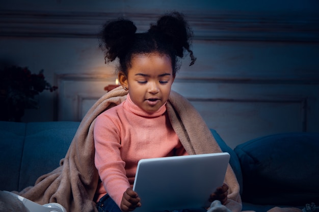 Feliz niña afroamericana durante la videollamada con dispositivos portátiles y domésticos, se ve encantada y feliz. Hablando con Santa antes de la víspera de Año Nuevo, su familia, tomando clases de preescolar en línea.