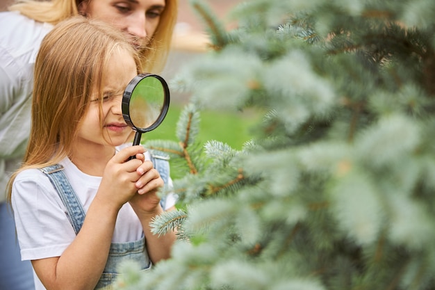 Feliz niña alegre mirando a través de la lupa al árbol de Navidad al aire libre con la madre