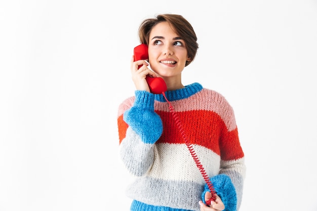 Feliz niña alegre vistiendo suéter que se encuentran aisladas en blanco, hablando por un teléfono fijo