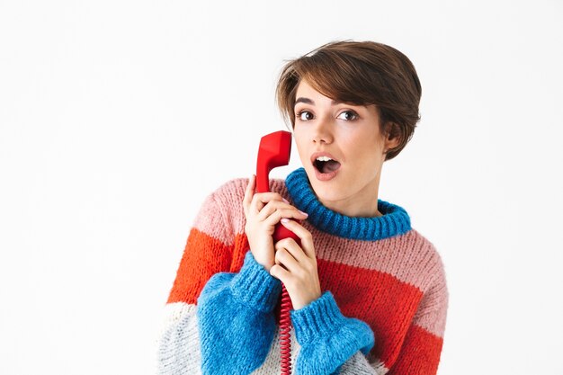 Feliz niña alegre vistiendo suéter que se encuentran aisladas en blanco, hablando por un teléfono fijo