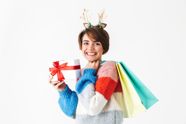 Feliz niña alegre vistiendo suéter que se encuentran aisladas en blanco, sosteniendo la caja actual, llevando bolsas de la compra.