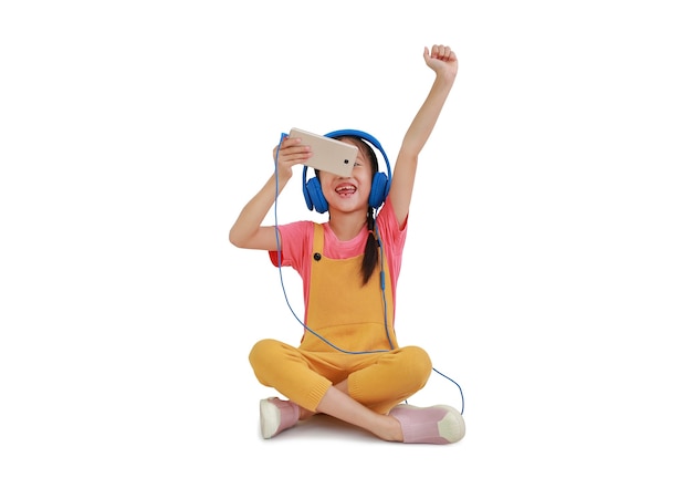 Feliz niña asiática con auriculares y disfrutar con smartphone sentado aislado en blanco