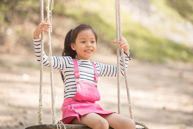 Feliz niña asiática divirtiéndose jugando en el patio de recreo en verano con una sonrisa y riendo saludable, Adorable niña divirtiéndose en un columpio en verano.