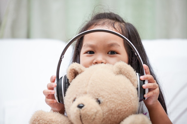 Feliz niña asiática jugando con su oso de peluche con auriculares en su habitación