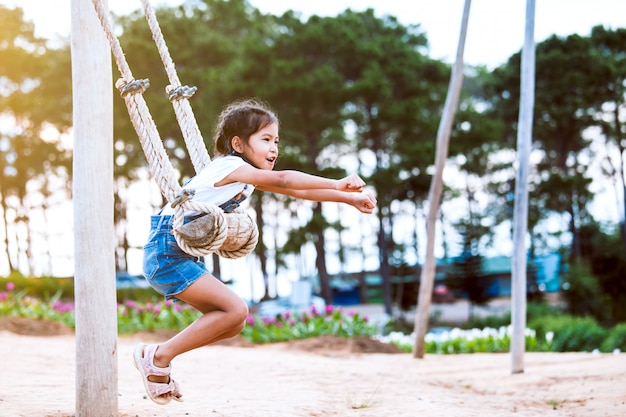 Feliz niña asiática niño divirtiéndose para jugar en columpios de madera en el patio de recreo con hermosa naturaleza