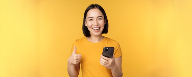 Feliz niña asiática sonriente sosteniendo el teléfono móvil y mostrando los pulgares hacia arriba recomendando la aplicación en el teléfono inteligente de pie sobre fondo amarillo