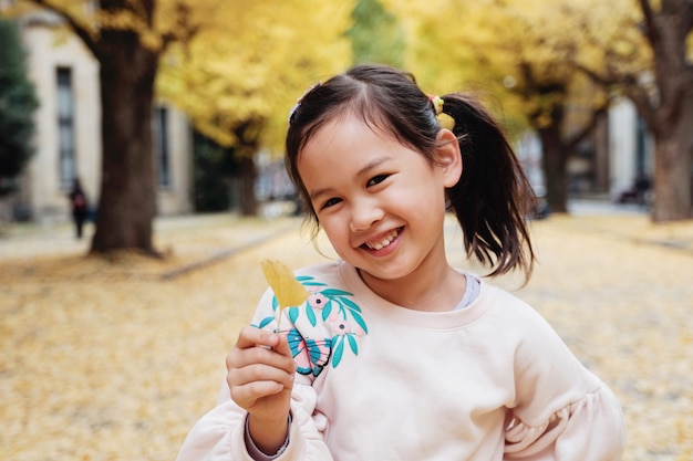 Feliz niña asiática sosteniendo una hoja de ginkgo amarillo y sonriendo en Tokio, Japón Otoño