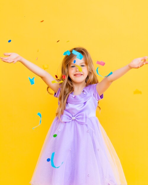 Feliz niña con cabello rubio y con un vestido azul atrapa confeti sobre un fondo amarillo, concepto de vacaciones