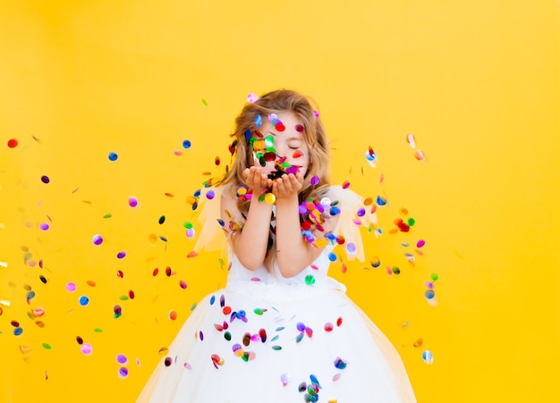 Feliz niña con cabello rubio y con un vestido blanco atrapa confeti sobre un fondo amarillo, concepto de vacaciones