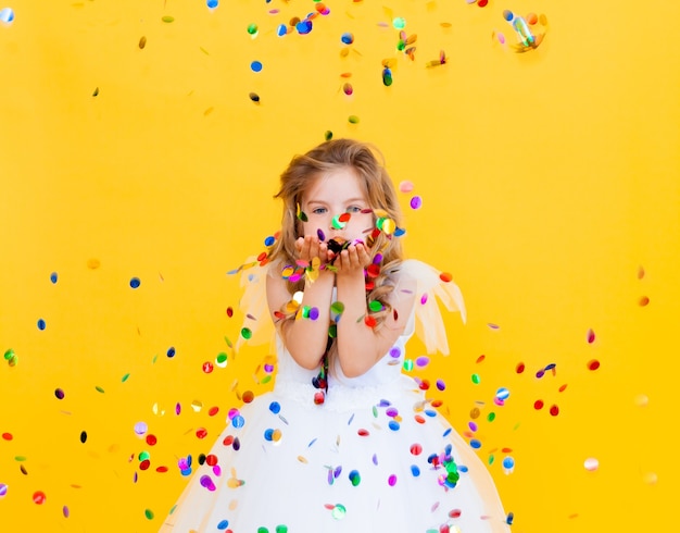 Feliz niña con cabello rubio y con un vestido blanco atrapa confeti sobre un fondo amarillo, concepto de vacaciones