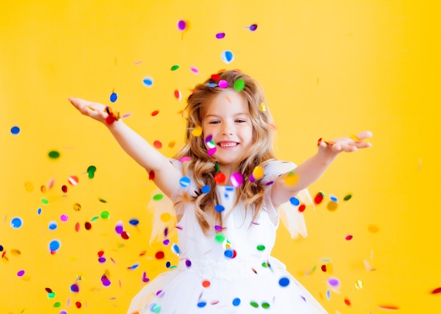 Feliz niña con cabello rubio y con un vestido blanco atrapa confeti sobre un fondo amarillo, concepto de vacaciones