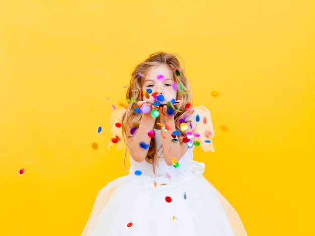 Feliz niña con cabello rubio y con un vestido blanco atrapa confeti sobre un fondo amarillo, concepto de vacaciones