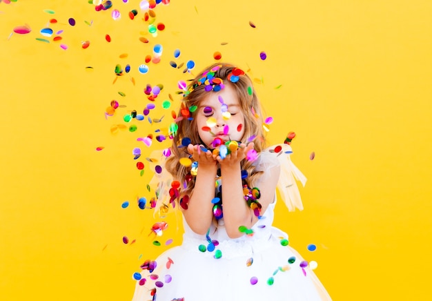 Feliz niña con cabello rubio y con un vestido blanco atrapa confeti sobre un fondo amarillo, concepto de vacaciones