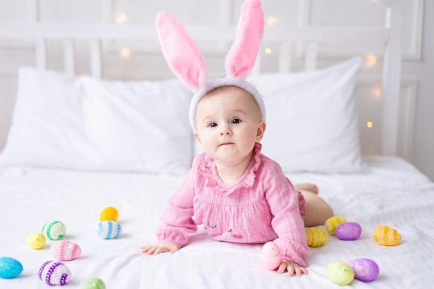 Feliz niña caucásica con un borde de orejas de conejo en la cabeza yace en la cama en casa en el dormitorio con coloridos huevos de Pascua bebé de Pascua