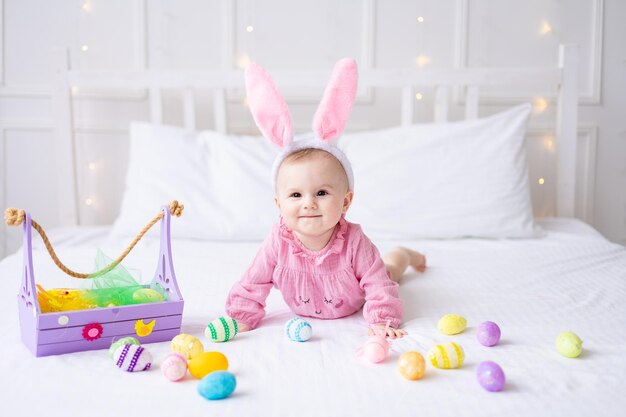 Feliz niña caucásica con un borde de orejas de conejo en la cabeza yace en la cama en casa en el dormitorio con coloridos huevos de Pascua bebé de Pascua