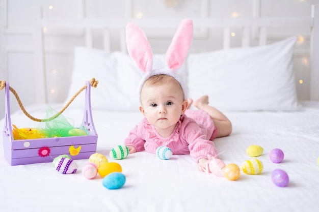 Feliz niña caucásica con un borde de orejas de conejo en la cabeza yace en la cama en casa en el dormitorio con coloridos huevos de Pascua bebé de Pascua