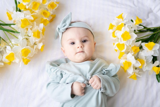 Una feliz niña caucásica con un traje verde está acostada de espaldas con flores amarillas de primavera en la cama de su casa en el dormitorio con ropa de cama blanca mirando a la cámara un bebé sano