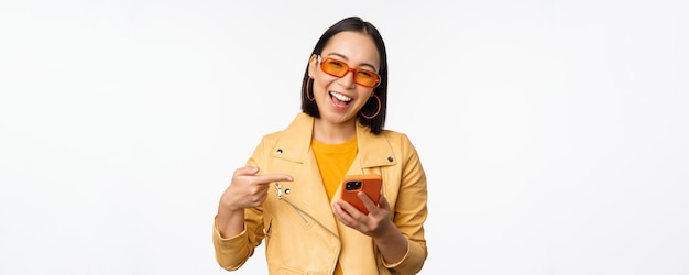 Feliz niña coreana sonriente con gafas de sol sosteniendo un teléfono inteligente apuntando a la pantalla del teléfono móvil de pie sobre fondo blanco Copiar espacio