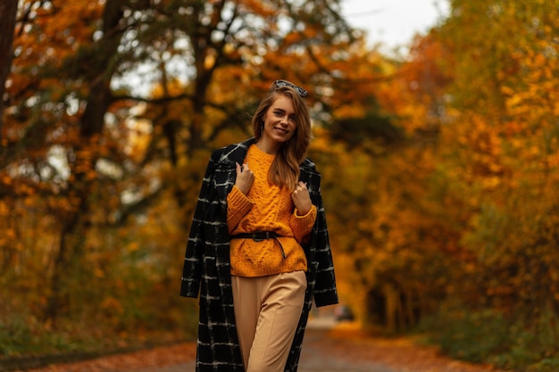 Feliz niña elegante con bonita sonrisa en ropa de colección otoño vintage con suéter de punto, abrigo negro camina al aire libre con hojas de naranja